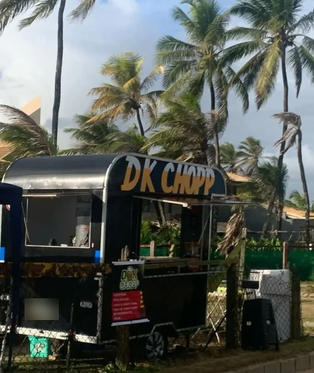 O trailer do DK Chopp: preto, com o letreiro DK CHOPP em amarelo, entre coqueiros e cercado por uma tela baixa.